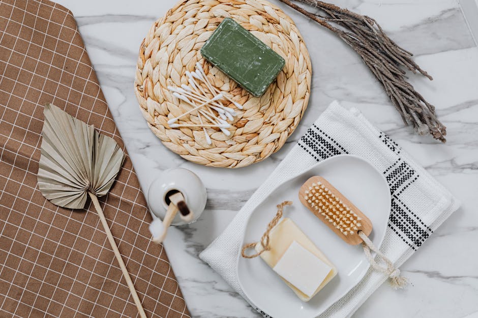 A flat-lay arrangement of bathroom or personal care items on a marble surface, including a round woven placemat with a green soap bar and white cotton swabs, a white ceramic vase with dried twigs, a f